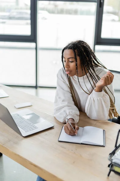 Afroamerikanische Geschäftsfrau schreibt auf Notizbuch neben Laptop auf Arbeitstisch — Stockfoto