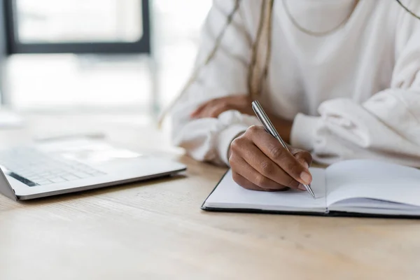 Vista recortada de la mujer de negocios afroamericana escribiendo en un cuaderno cerca de la computadora portátil en la oficina - foto de stock