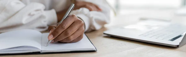 Vista recortada de la mujer de negocios afroamericana escribiendo en el cuaderno cerca de la computadora portátil en la mesa, bandera - foto de stock