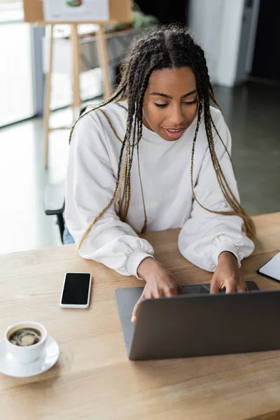 Femme d'affaires afro-américaine souriante utilisant un ordinateur portable près du café et un smartphone au bureau — Photo de stock