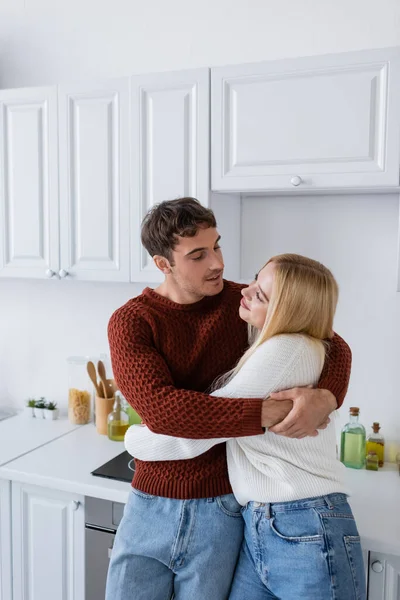 Jovem em camisola vermelha abraçando alegre namorada loira na cozinha — Fotografia de Stock