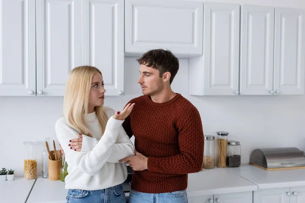 Jovem homem em camisola vermelha olhando para namorada loira na cozinha — Fotografia de Stock