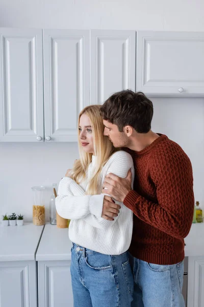 Tendre homme en pull rouge étreignant jeune copine blonde dans la cuisine — Photo de stock