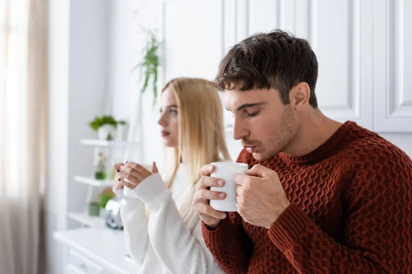 Junger Mann im Strickpullover hält Tasse in der Hand und bläst beim Aufwärmen neben verschwommener Freundin zum Tee — Stockfoto