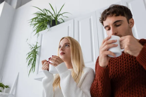 Vista basso angolo di giovane donna in maglia maglione tenendo tazza di tè vicino fidanzato offuscata — Foto stock