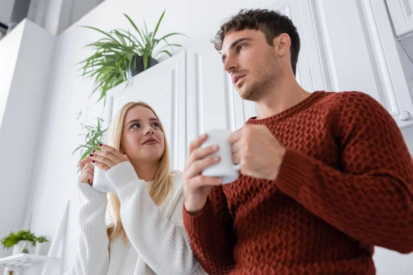 Low angle view of young woman in knitted sweater holding cup of tea while looking at boyfriend — Stock Photo