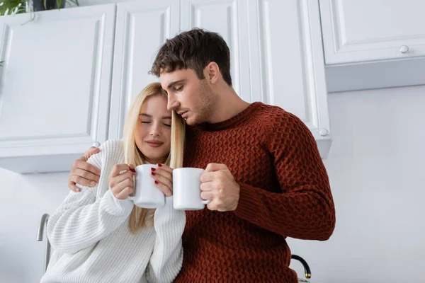 Pareja joven en suéteres de punto abrazando y sosteniendo tazas con té mientras se calienta en invierno - foto de stock