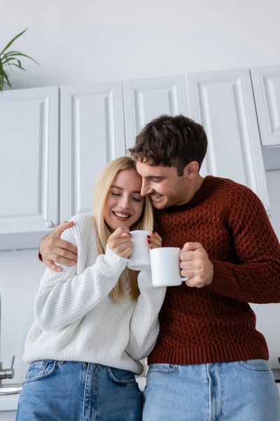Feliz pareja joven en suéteres de punto abrazando y sosteniendo tazas con té mientras se calienta en invierno - foto de stock