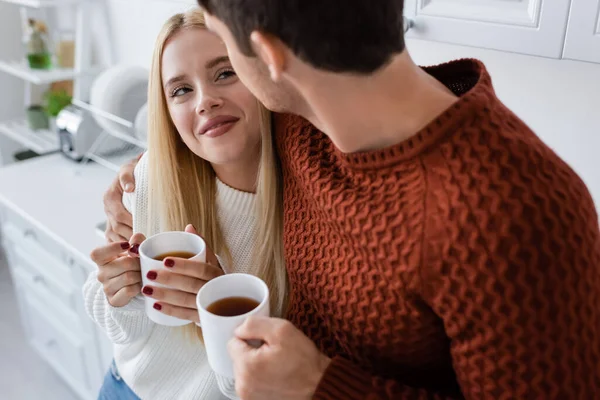 Cheerful young couple in knitted sweaters hugging and holding cups with tea while warming up in winter — Stock Photo