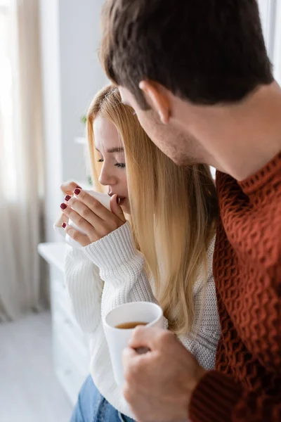 Young woman in knitted sweater drinking tea while warming up near boyfriend — Stock Photo