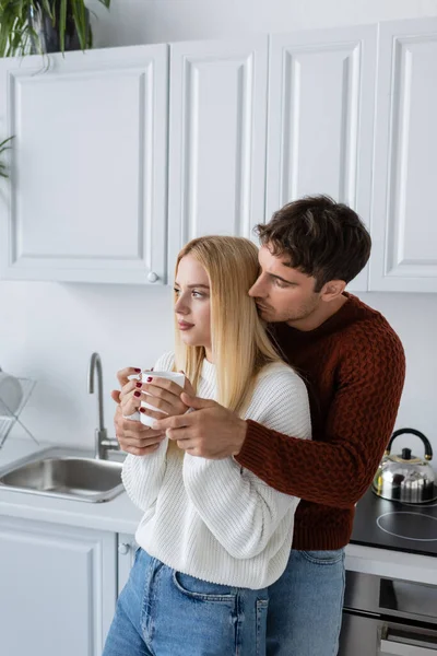 Молодий чоловік в трикотажному светрі обіймає блондинку з чашкою чаю взимку — стокове фото