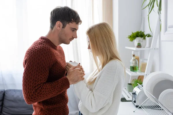 Vista lateral del hombre joven en suéter rojo y mujer rubia calentándose mientras sostiene la taza de té - foto de stock