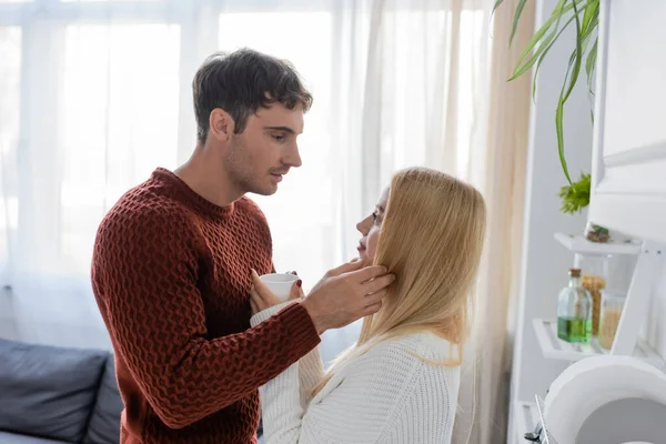 Vista lateral del hombre en suéter rojo tocando la cara de la mujer rubia sosteniendo la taza de té - foto de stock