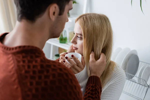 Hombre joven en suéter rojo acariciando el pelo de la mujer rubia sosteniendo la taza de té - foto de stock