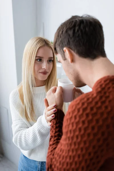 Blonde woman looking at boyfriend in red sweater holding cup and drinking tea — Stock Photo