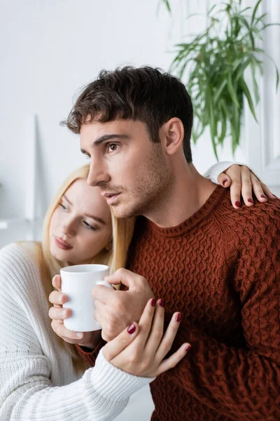 Mujer rubia abrazando novio en suéter rojo con taza de té - foto de stock