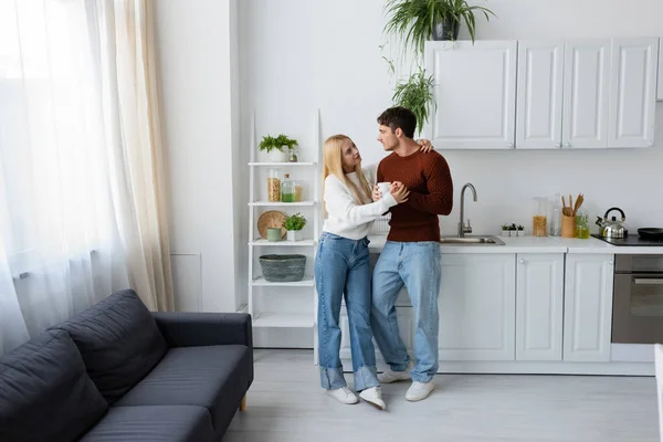 Full length of young and blonde woman hugging boyfriend in red sweater with cup of tea — Stock Photo