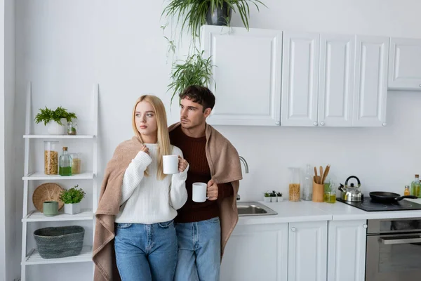 Jovem casal em camisolas de pé coberto de cobertor e segurando xícaras de chá — Fotografia de Stock
