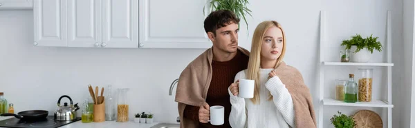 Молода пара в светрах стоїть в ковдрі і тримає чашки чаю, банер — стокове фото