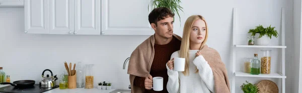 Joven pareja en suéteres de pie cubierto de manta y la celebración de tazas de té, pancarta - foto de stock