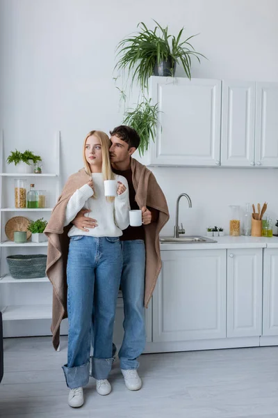 Young couple covered in blanket hugging and holding cups of tea — Stock Photo