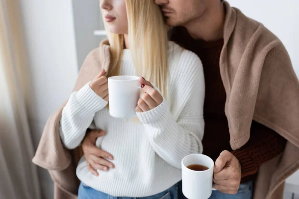 Cropped view of young couple covered in blanket hugging while holding cups of tea at home — Stock Photo