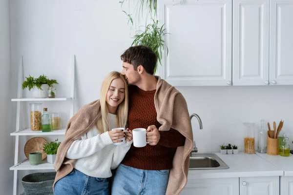 Joyeux jeune couple couvert de couverture embrassant tout en tenant des tasses de thé à la maison — Photo de stock