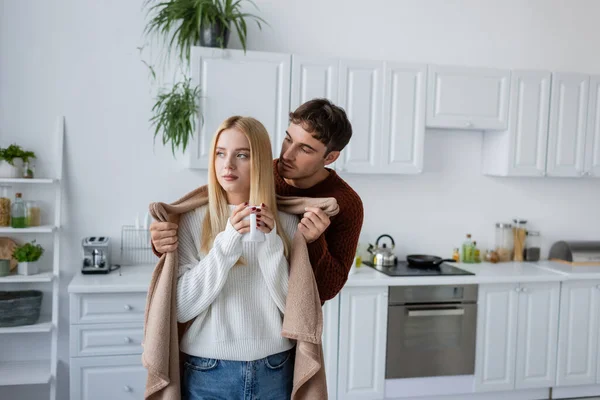 Дбайливий чоловік тримає ковдру біля дівчини в светрі з чашкою чаю — стокове фото