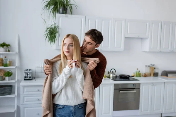 Caring man holding blanket near girlfriend in sweater with cup of tea — Stock Photo