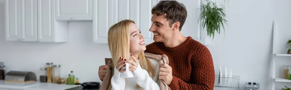 Hombre cariñoso sosteniendo manta cerca de novia feliz en suéter con taza de té, pancarta - foto de stock