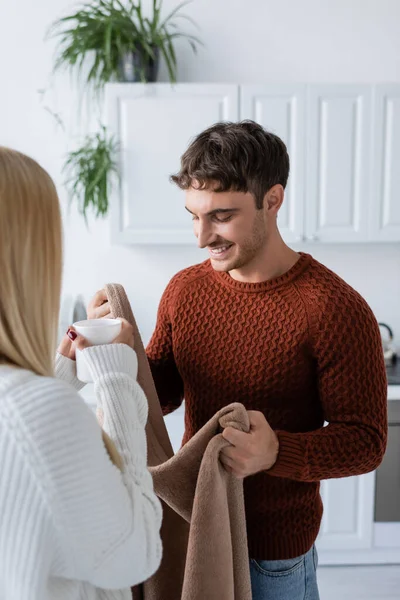 Hombre alegre sosteniendo manta cerca de novia en suéter con taza de té - foto de stock