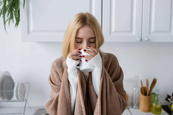 Молодая блондинка в свитере и одеяле пьет чай из чашки — стоковое фото