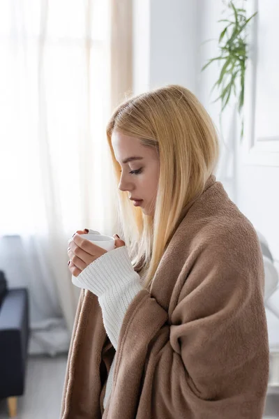 Jeune femme blonde en pull blanc et couverture regardant une tasse de thé — Photo de stock