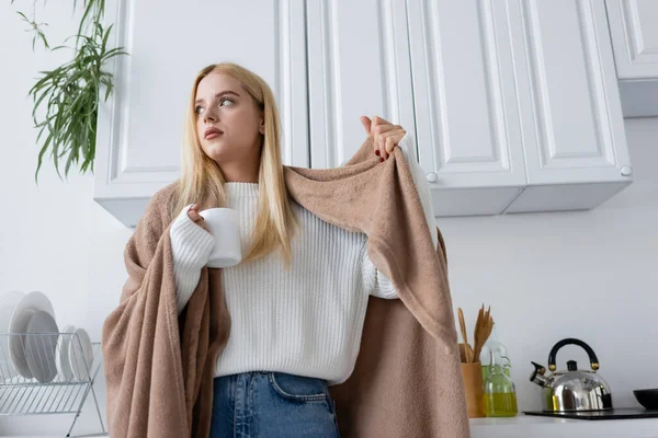 Low angle view of young blonde woman in white sweater and blanket holding cup of tea — Stock Photo