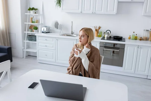 Mujer rubia cubierta de manta sosteniendo taza de té cerca de gadgets en la mesa - foto de stock