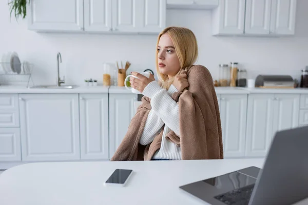 Blonde woman covered in blanket holding cup of tea near devices on table — Stock Photo