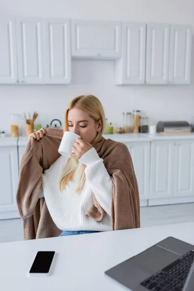 Donna bionda coperta di coperta che beve tè vicino ai gadget sul tavolo — Foto stock