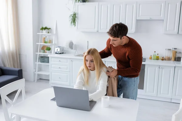 Uomo premuroso in maglione che tiene la coperta vicino alla ragazza che lavora da casa sul computer portatile — Foto stock