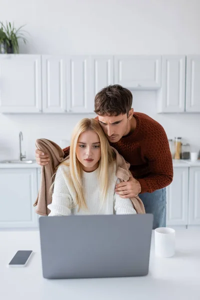 Caring boyfriend in sweater holding blanket near tensed girlfriend working from home on laptop — Stock Photo