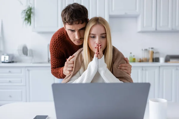 Hombre en suéter calmando novia preocupada mirando a la computadora portátil blanca trabajando remotamente en casa - foto de stock