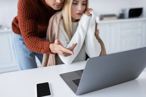 Homme en pull pointant vers ordinateur portable près petite amie blonde et smartphone sur le bureau — Photo de stock