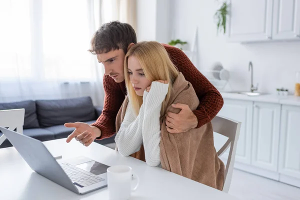 Homme en pull pointant vers ordinateur portable près de jeune copine et tasse de thé sur la table — Photo de stock