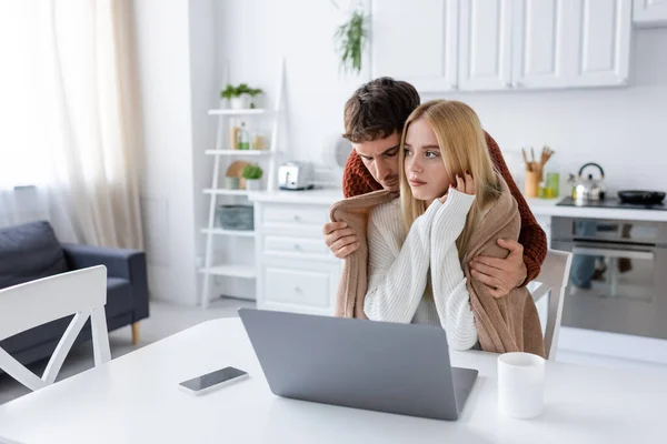 Premuroso fidanzato in maglione abbracciare fidanzata in coperta seduto vicino gadget sul tavolo — Foto stock