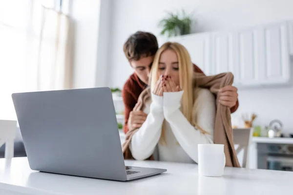 Portátil y taza en el escritorio cerca de novio cariñoso en suéter que cubre novia rubia congelante con manta - foto de stock