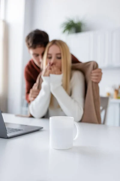 Taza de té en el escritorio cerca de novio cariñoso en suéter que cubre novia rubia congelante con manta - foto de stock