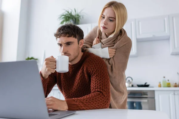 Mujer rubia cubierta de manta de pie detrás de novio beber té mientras trabaja desde casa - foto de stock