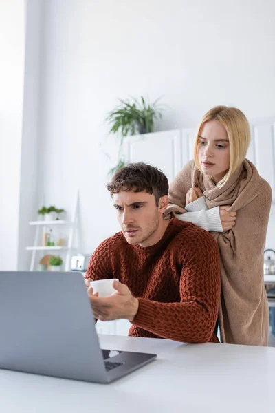 Mujer rubia cubierta de manta de pie detrás de novio usando el ordenador portátil mientras trabaja desde casa - foto de stock