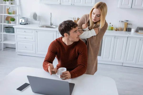 Femme blonde couverte de couverture debout derrière petit ami travaillant de la maison près de gadgets — Photo de stock