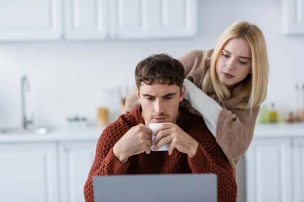 Femme blonde couverte de couverture debout derrière petit ami avec une tasse de thé travaillant à la maison — Photo de stock