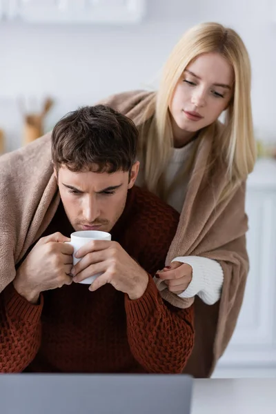 Junge Frau in Decke gehüllt steht hinter Freund mit Tasse Tee und arbeitet von zu Hause aus — Stockfoto